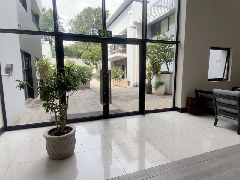 A bright, modern commercial entrance lobby featuring large glass panels, double doors, and polished tiled flooring, providing a clear view of the outdoor courtyard.