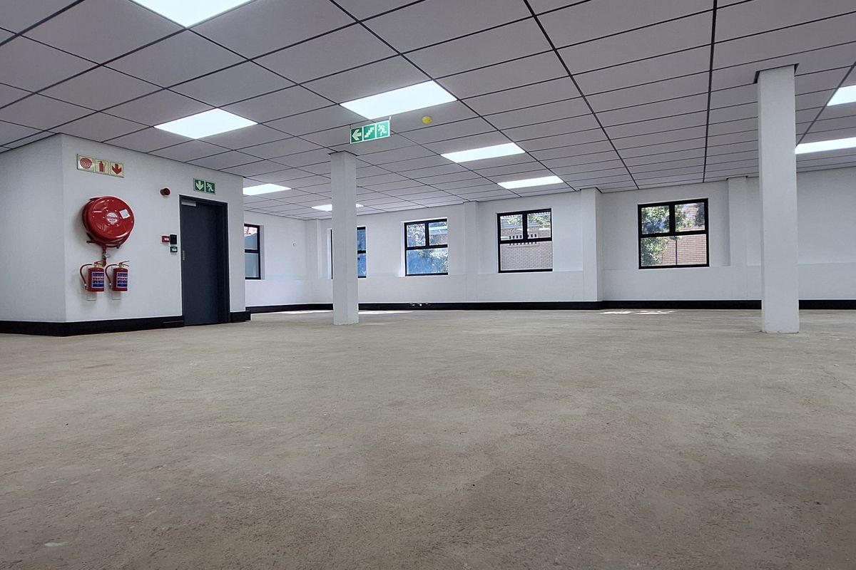 A spacious, open-plan office area featuring a suspended ceiling with recessed lighting, structural columns, and essential fire safety equipment.