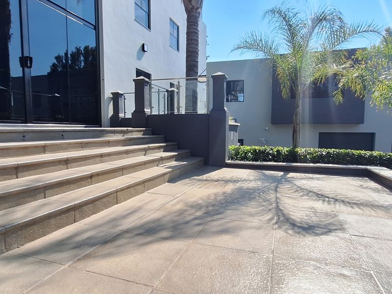A spacious, modern tiled patio area featuring stone steps leading to large glass sliding doors and a contemporary glass balustrade.