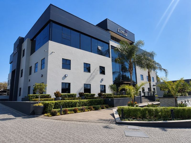Modern multi-story office building featuring a contemporary facade, glass curtain walls, paved driveway, and professionally landscaped gardens with a water feature.