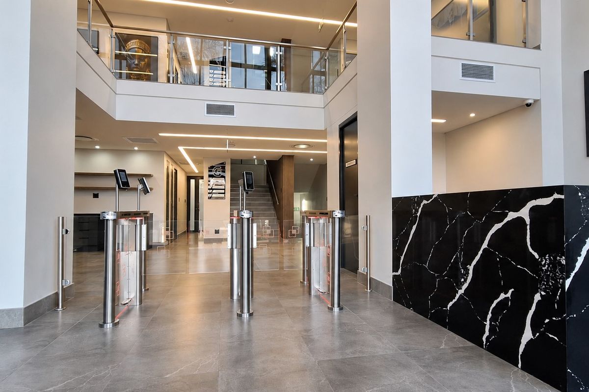 Modern office building entrance lobby featuring security turnstiles, a marble-finish reception desk, and an open-plan mezzanine level with glass balustrades.