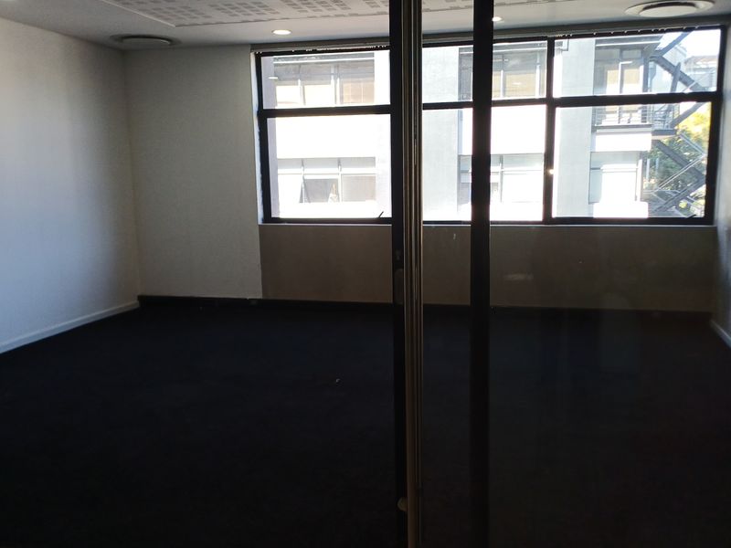 An empty office space featuring large windows, carpeted flooring, recessed lighting, and a glass partition door, offering a clean and professional work environment.