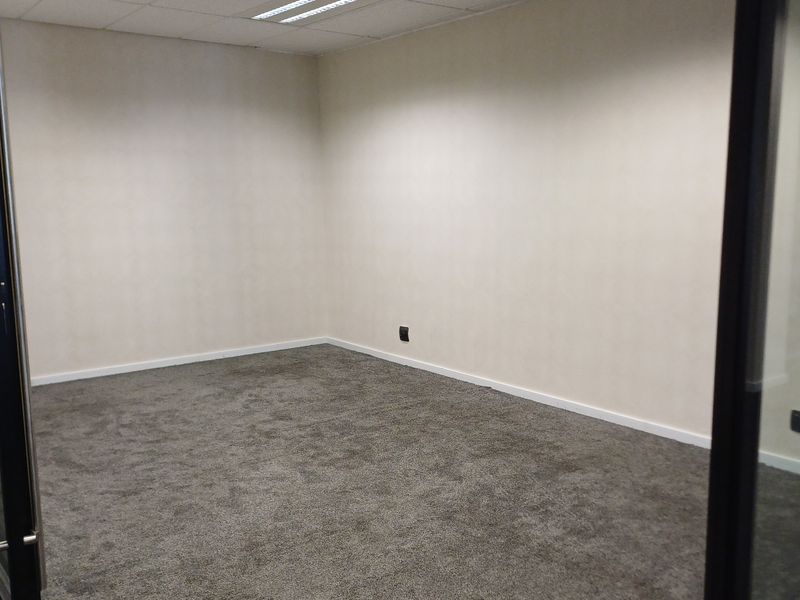 A clean, empty office space featuring grey carpeting, neutral walls, a suspended ceiling with recessed lighting, and modern glass partition walls.