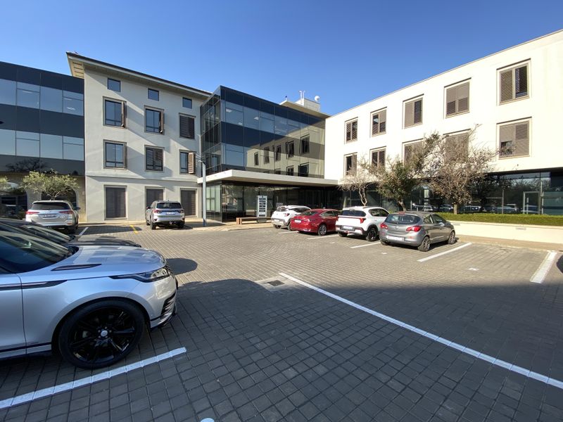 Modern office building exterior featuring a glass facade, paved parking area, and integrated window shutters, presenting a professional corporate appearance.