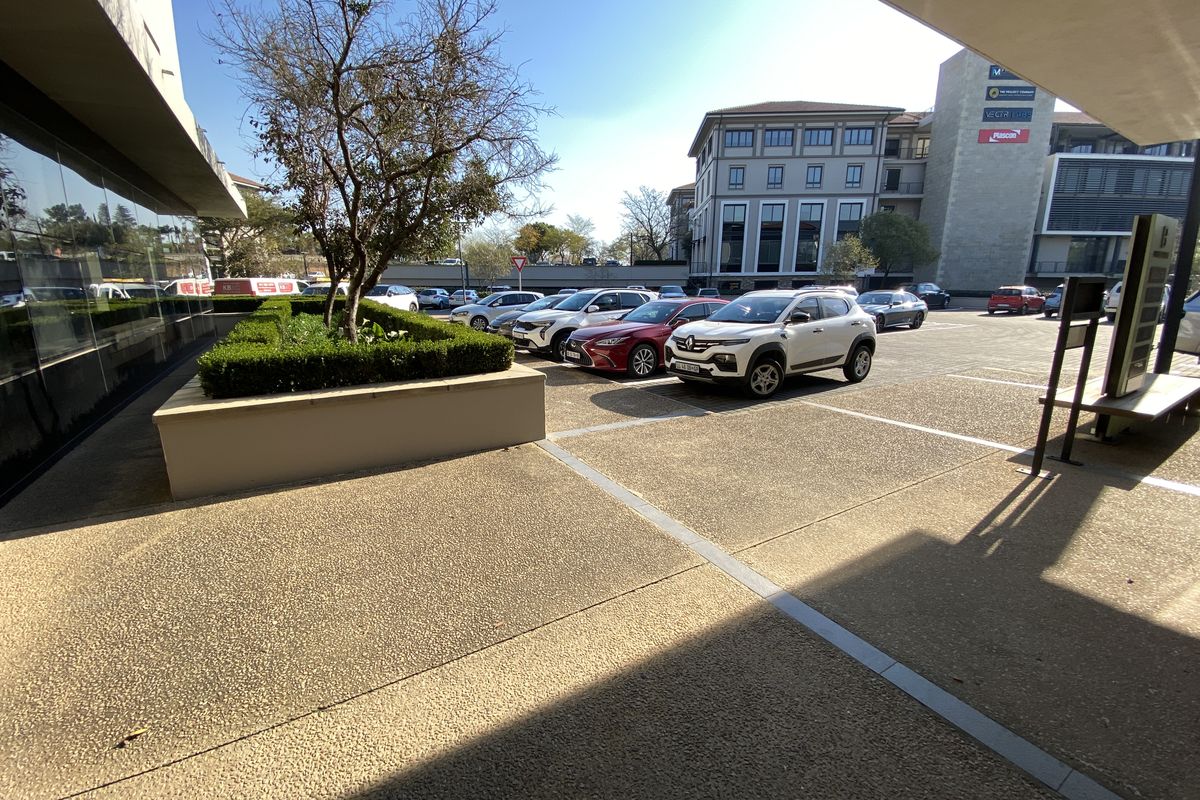 A view of the office building exterior featuring a paved parking area, landscaped planter box, and modern glass facade.