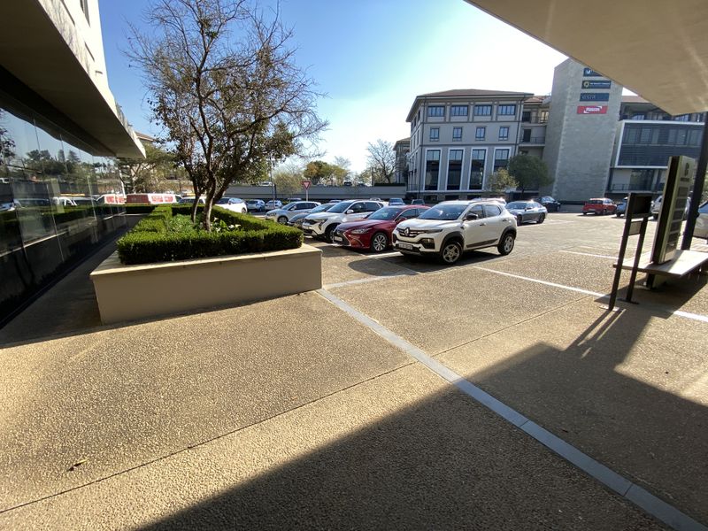 A view of the office building exterior featuring a paved parking area, landscaped planter box, and modern glass facade.
