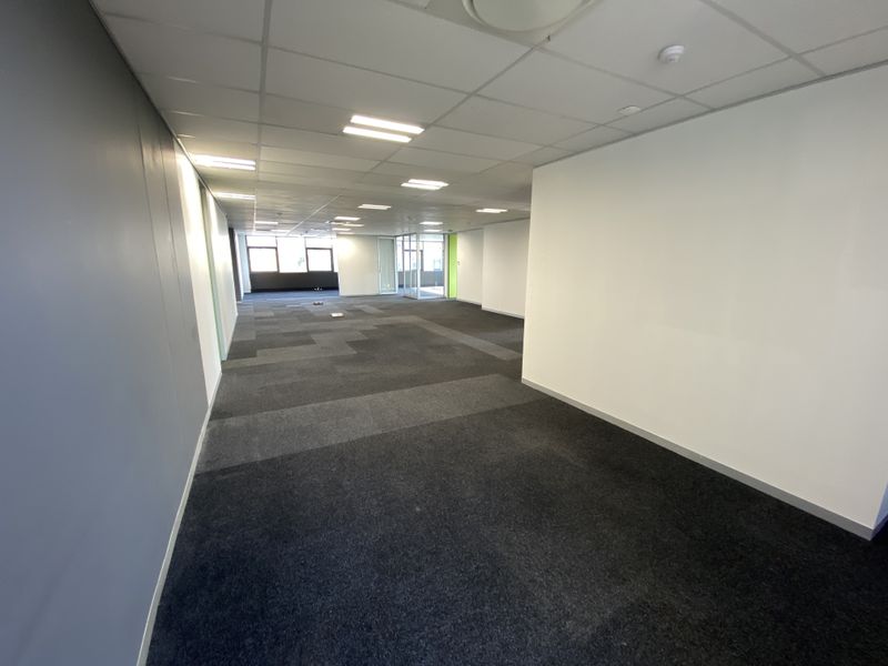 A spacious, open-plan office area featuring neutral carpet tiles, a suspended ceiling with recessed lighting, and a glass partition.