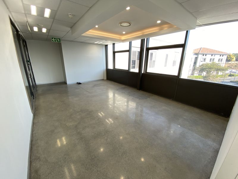 A bright, empty office space featuring polished concrete floors, a modern suspended ceiling with recessed lighting, and large windows providing ample natural light.