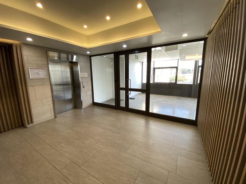 Modern office building lobby featuring a stainless steel elevator, tiled flooring, timber slat wall accents, and glass partitions leading to an open office area.