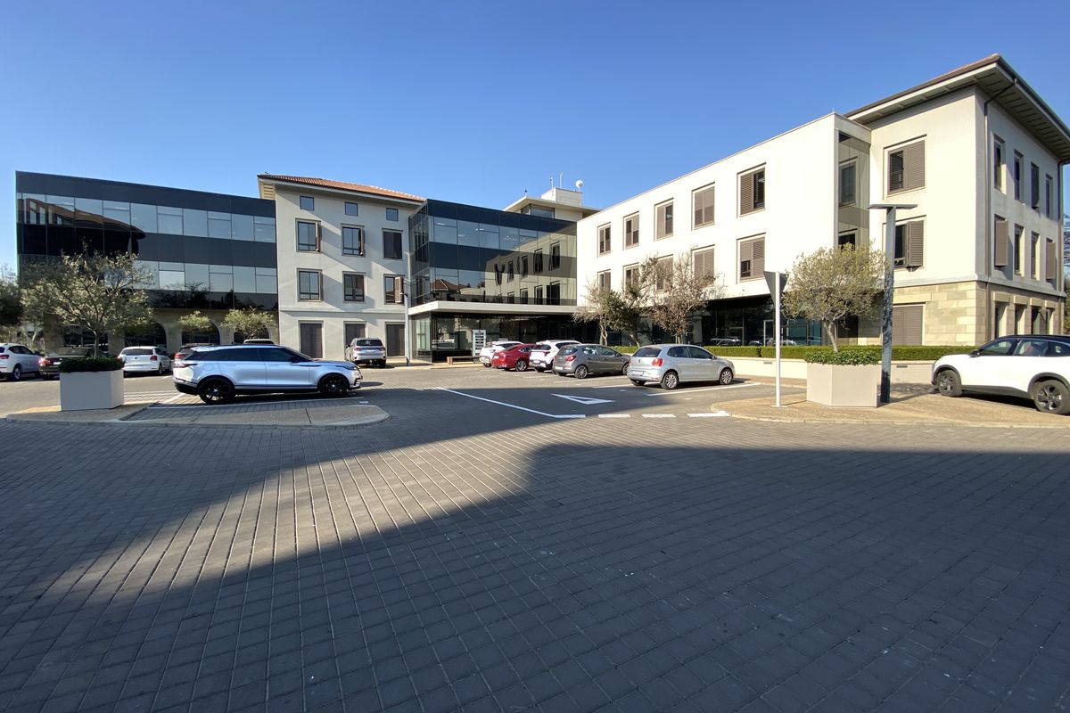 Modern multi-story office building featuring a combination of glass curtain walls, stone cladding, and paved parking area.
