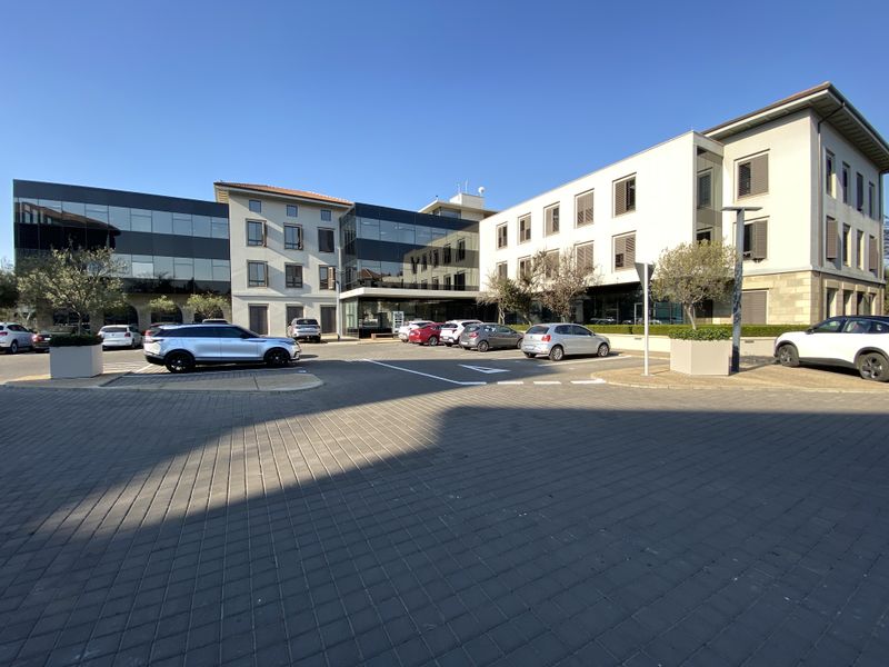 Modern multi-story office building featuring a combination of glass curtain walls, stone cladding, and paved parking area.