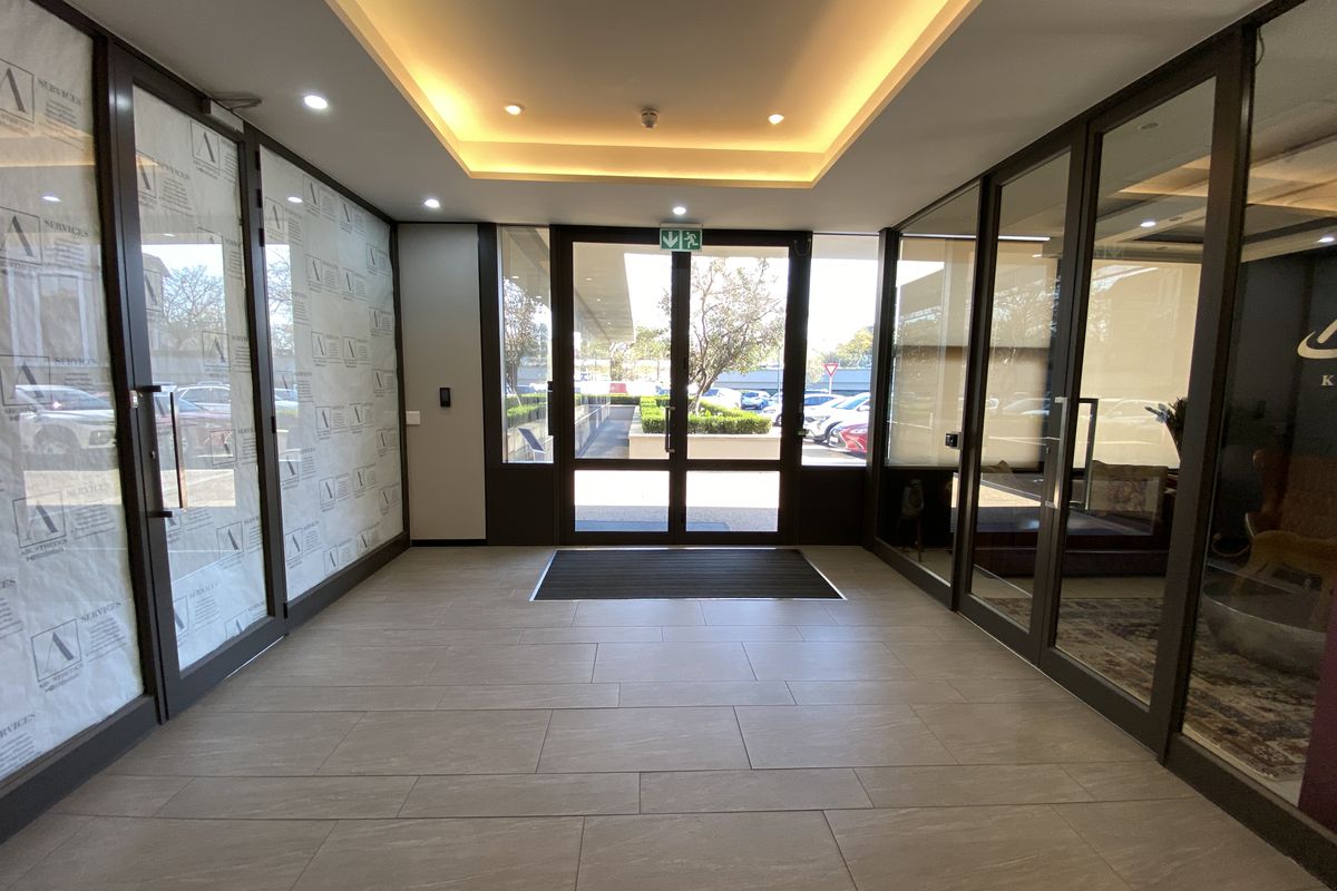 Modern office building entrance lobby featuring tiled flooring, recessed ceiling lighting, and glass partition walls providing a bright and professional welcome area.