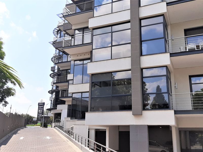 Modern multi-story office building featuring a glass facade, private balconies with stainless steel railings, and a paved driveway leading to undercover parking.
