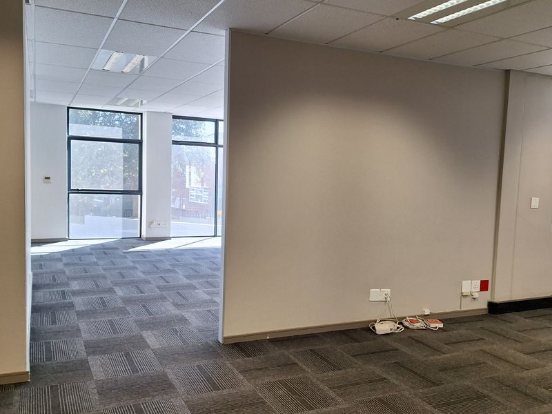 A bright, open-plan office space featuring neutral walls, carpet tile flooring, suspended ceilings with recessed lighting, and large windows providing natural light.