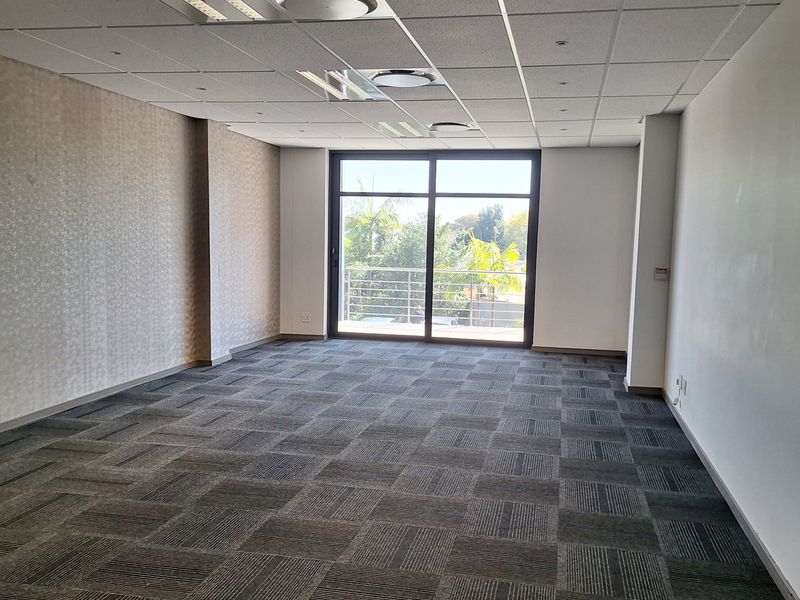 A bright, empty office space featuring carpet tiles, a suspended ceiling with recessed lighting, and a large sliding glass door leading to a balcony.