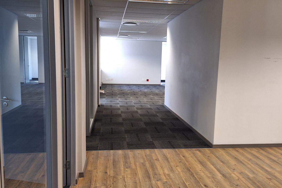 A vacant office space featuring a mix of laminate and carpet flooring, glass partitions, and a suspended ceiling with recessed lighting.