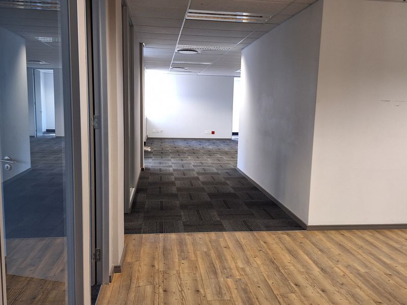 A vacant office space featuring a mix of laminate and carpet flooring, glass partitions, and a suspended ceiling with recessed lighting.