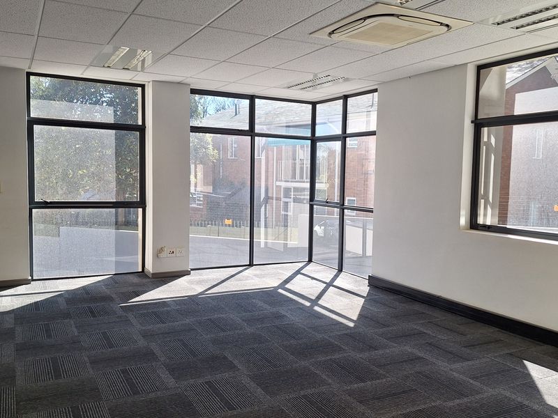 A bright, vacant office space featuring large windows, carpet tiles, a suspended ceiling with recessed lighting, and a ceiling-mounted air conditioning unit.