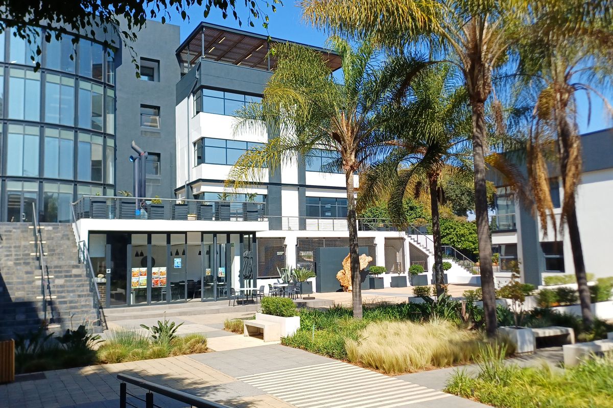 A modern multi-story office building featuring a landscaped courtyard, paved walkways, and a glass-fronted ground floor entrance with an elevated terrace.