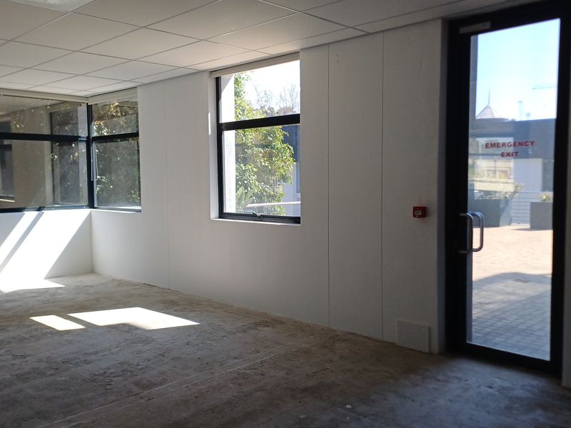 An empty office space featuring large windows, a suspended ceiling, and a dedicated emergency exit door with direct access to the exterior.