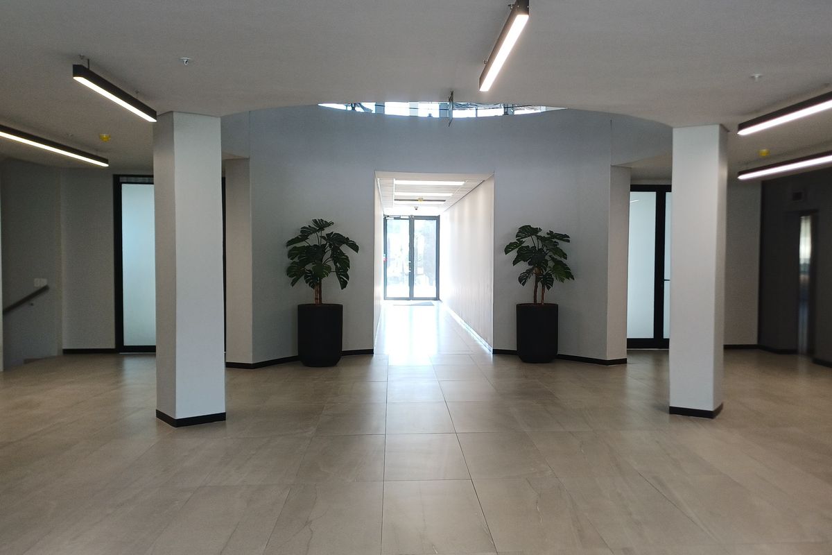 A spacious and modern office building entrance lobby featuring tiled flooring, structural columns, and bright recessed ceiling lighting.