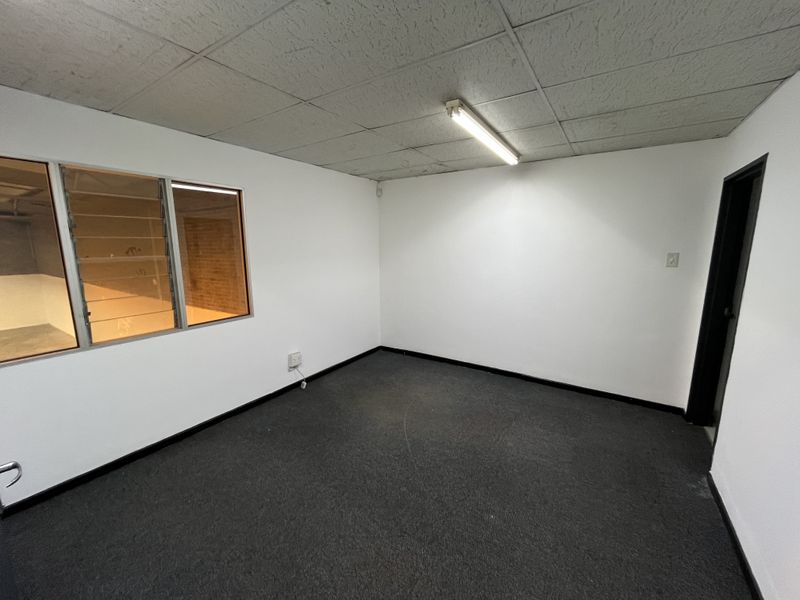A vacant office space featuring a suspended ceiling, fluorescent lighting, carpeted flooring, and a louvre window looking into an adjacent area.