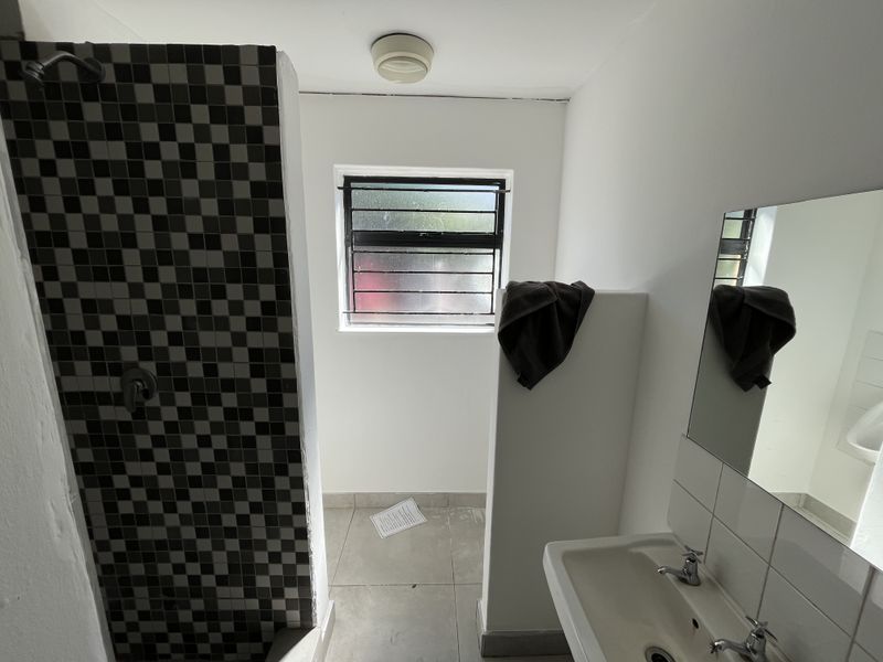 A functional bathroom featuring a tiled shower enclosure, a wall-mounted basin, and a window with security bars, providing basic utility and natural light.