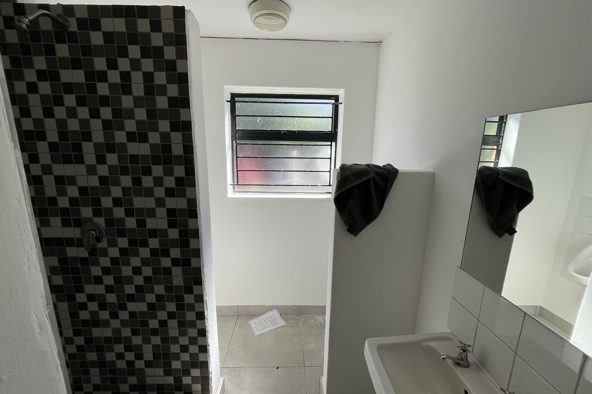A functional bathroom featuring a tiled shower enclosure, a wall-mounted basin, and a window with security bars, providing basic utility and natural light.