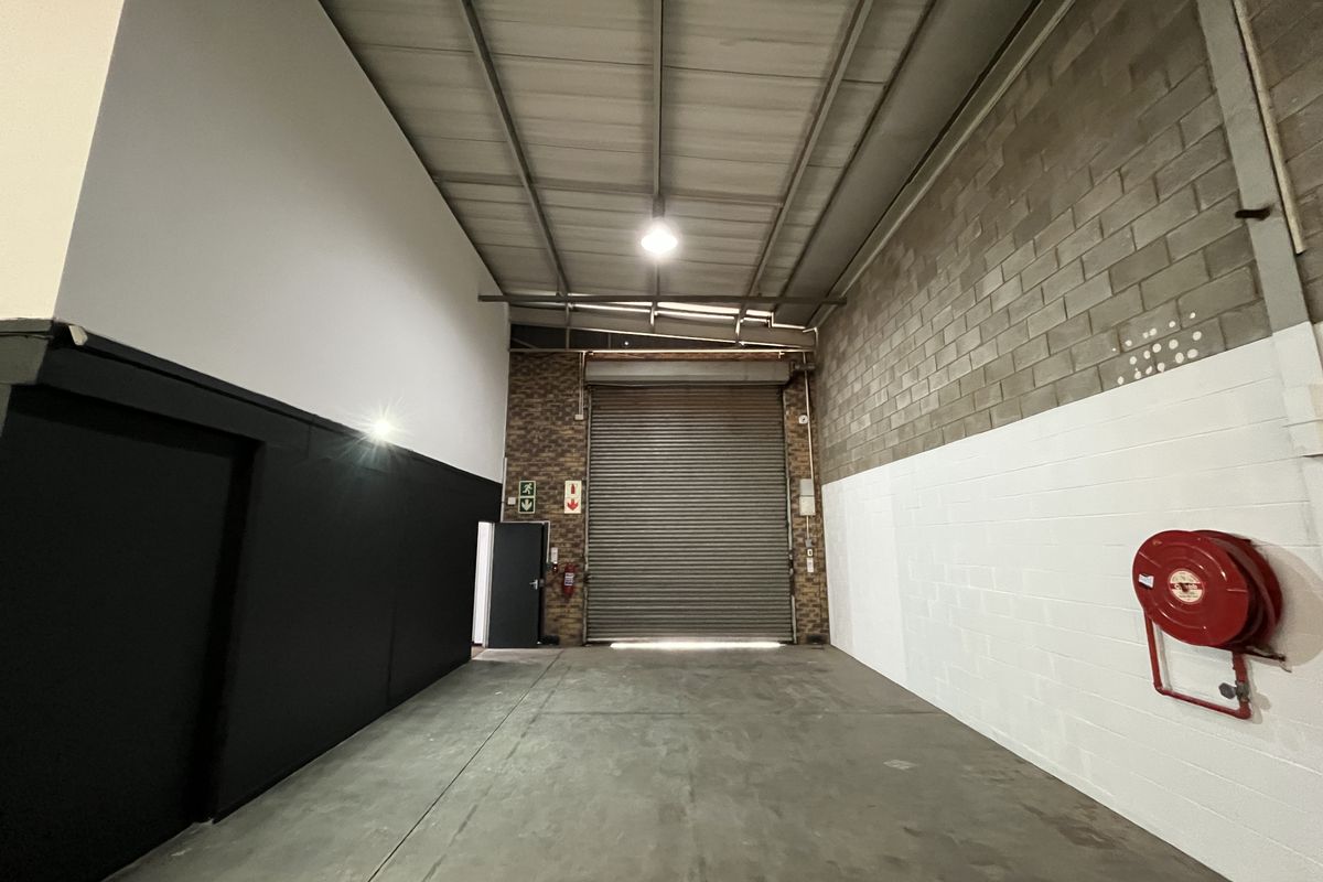 Spacious industrial warehouse unit featuring high ceilings with exposed steel trusses, concrete flooring, and a large roller shutter door for easy access.