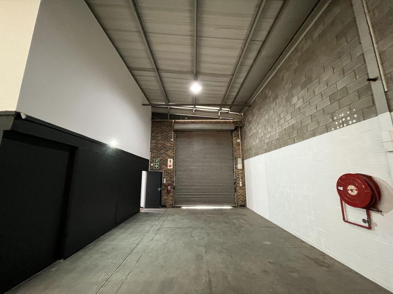 Spacious industrial warehouse unit featuring high ceilings with exposed steel trusses, concrete flooring, and a large roller shutter door for easy access.