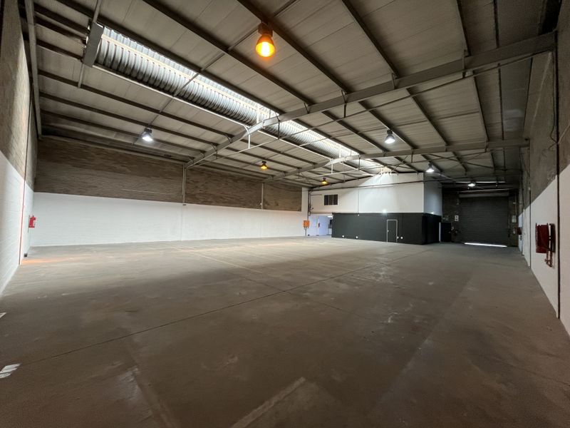 Spacious industrial warehouse featuring high ceilings with exposed steel trusses, skylights, concrete flooring, and a large roller shutter door for easy access.