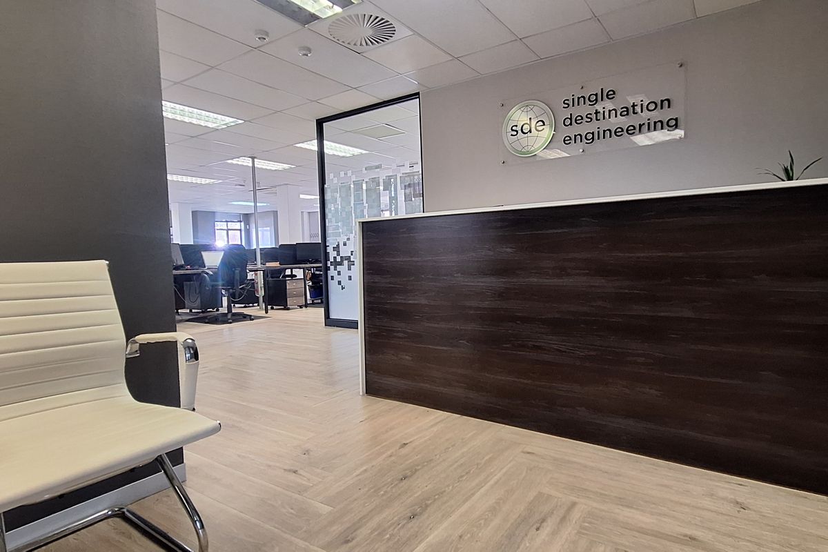 Professional office reception area featuring a dark wood-finish reception desk, modern laminate flooring, and a glass partition leading into the main workspace.
