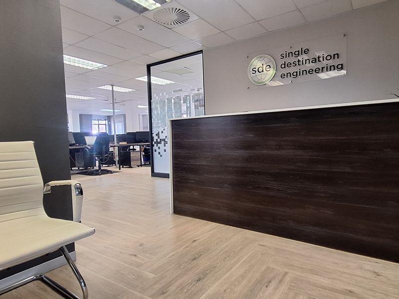 Professional office reception area featuring a dark wood-finish reception desk, modern laminate flooring, and a glass partition leading into the main workspace.