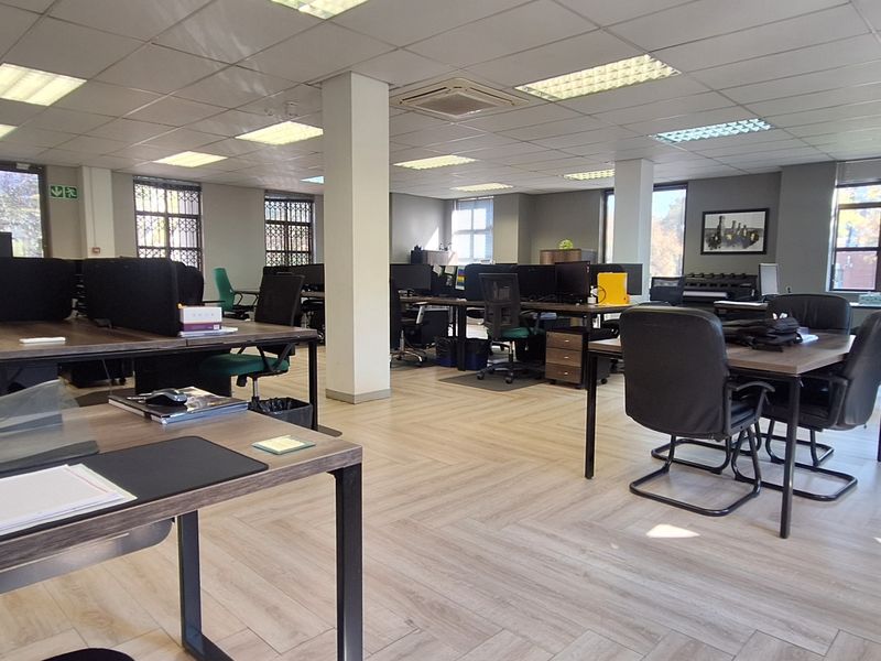 Spacious open-plan office area featuring laminate flooring, suspended ceilings with recessed lighting, and large windows with security bars.