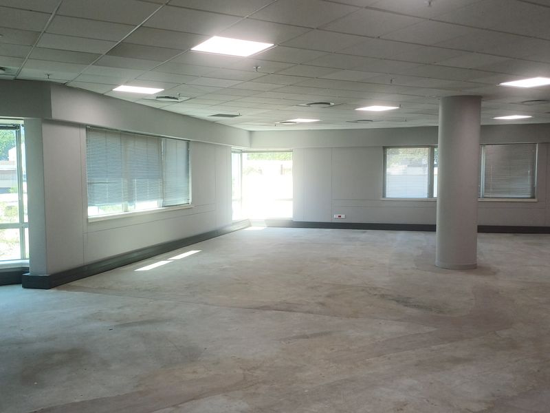 A spacious, vacant open-plan office area featuring a suspended ceiling, recessed lighting, structural columns, and large windows with venetian blinds.