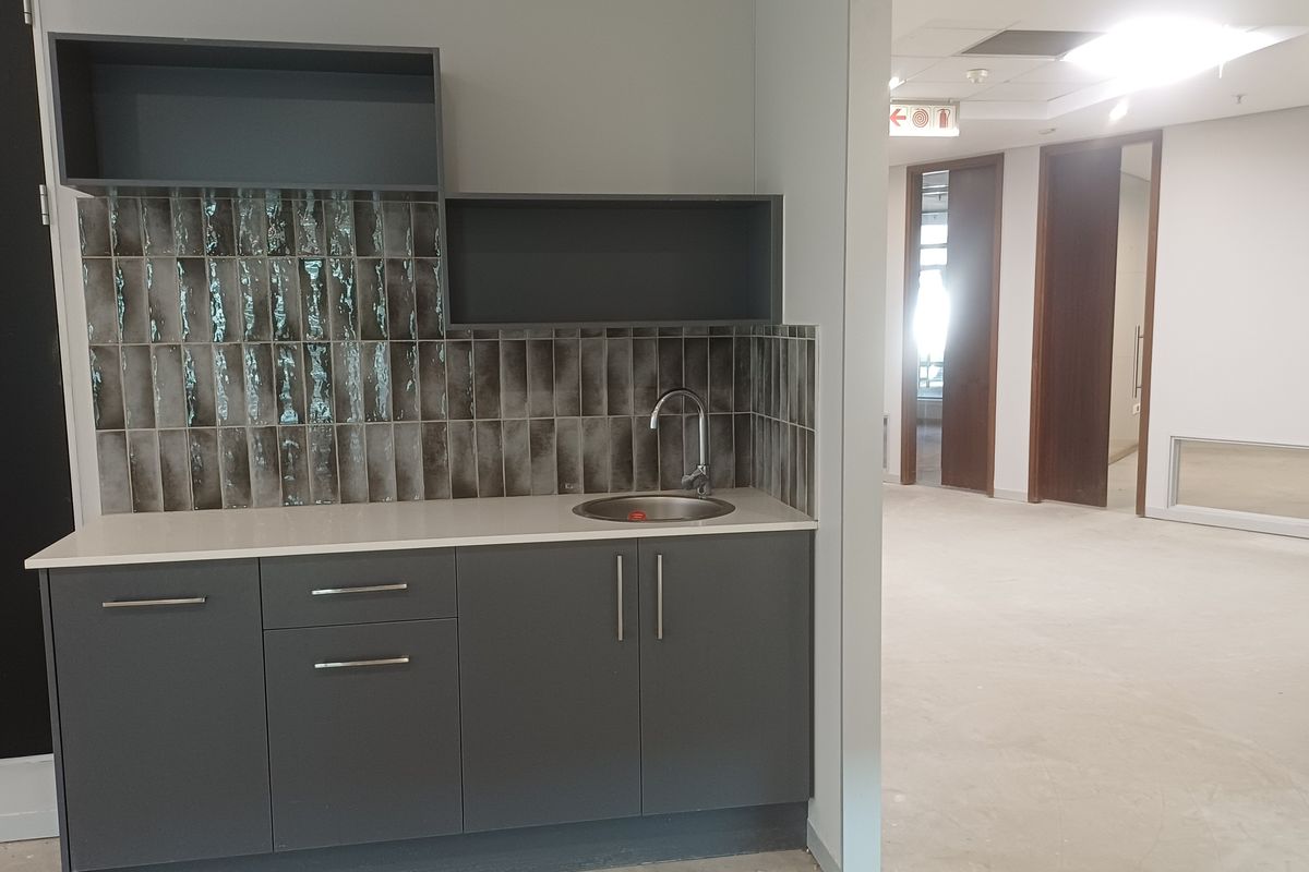 A modern office kitchenette featuring dark grey cabinetry, a sleek sink, and a textured tiled backsplash, adjacent to an open-plan office area.