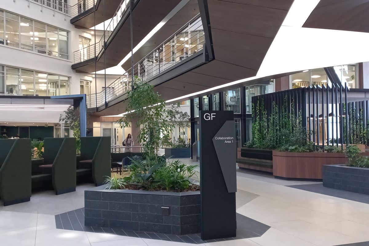 Modern open-plan office atrium featuring multi-level mezzanine walkways, tiled flooring, integrated planters, and contemporary architectural design elements.