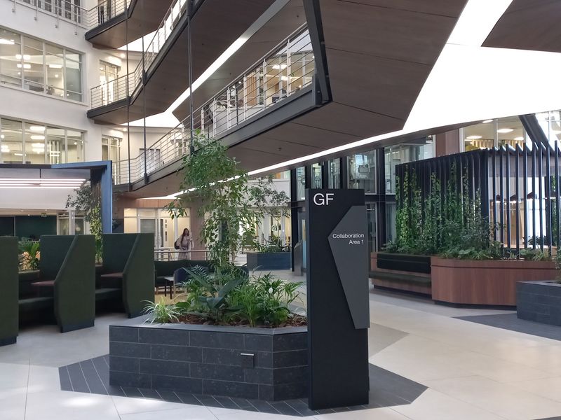 Modern open-plan office atrium featuring multi-level mezzanine walkways, tiled flooring, integrated planters, and contemporary architectural design elements.