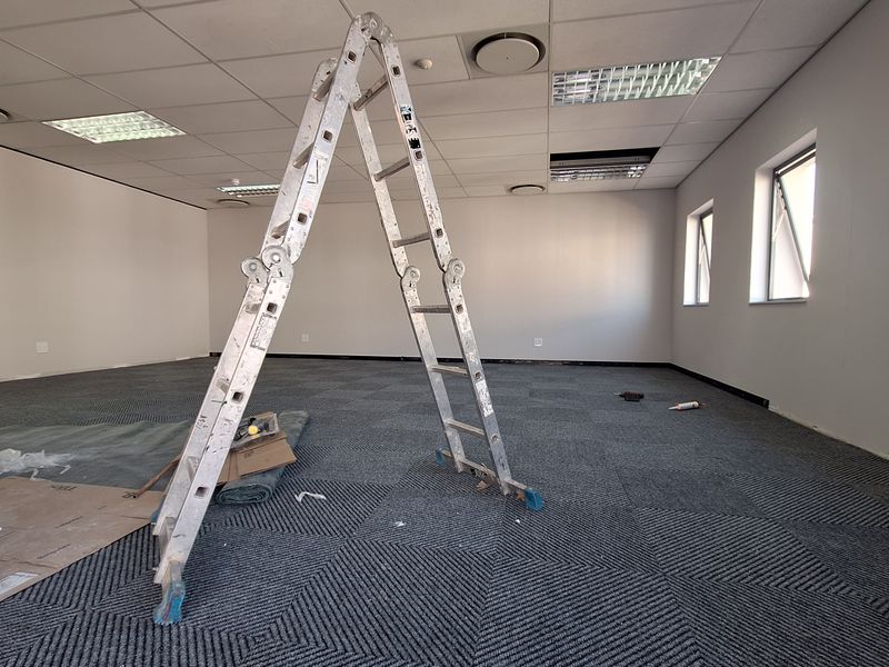 An open-plan office space featuring carpet tiles, a suspended ceiling with recessed lighting, and multiple windows providing natural light.