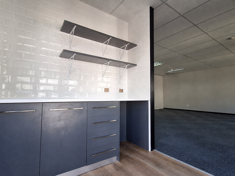 A modern office kitchenette featuring dark grey cabinetry, white subway tile backsplash, floating shelves, and an adjacent open-plan office area with carpeted flooring.