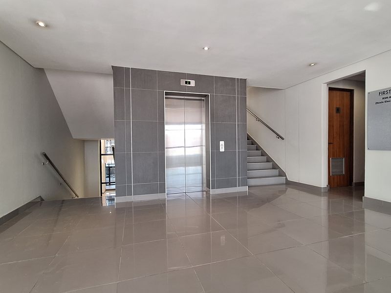 A clean and modern office building lobby featuring a central elevator, staircase access, polished tile flooring, and professional directory signage.