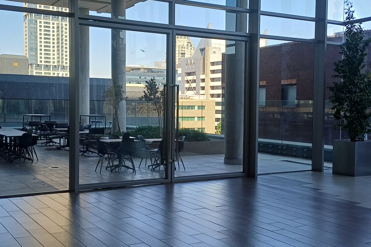 A spacious outdoor terrace featuring tiled flooring, concrete structural columns, and a glass balustrade, accessible through large floor-to-ceiling glass sliding doors.