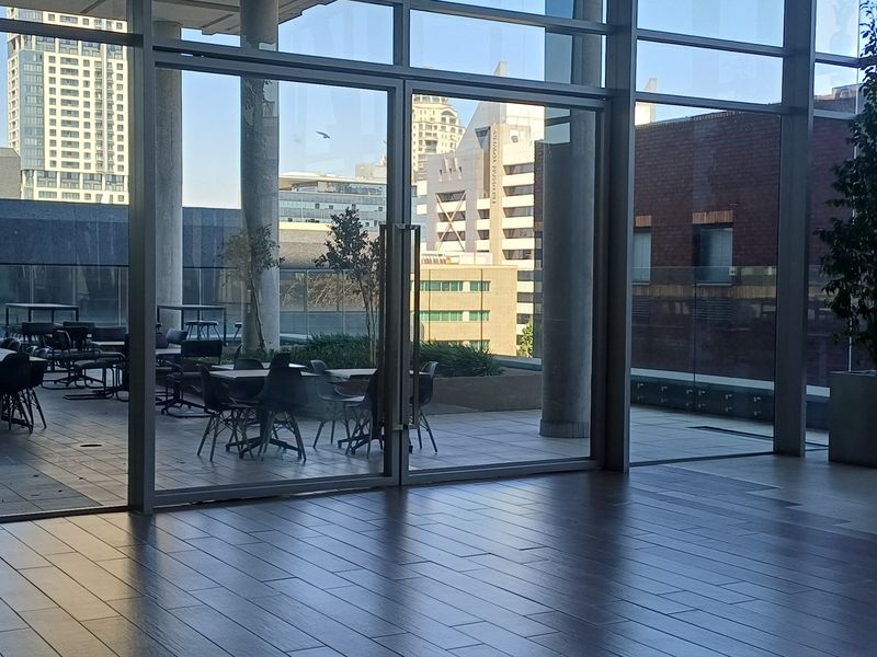 A spacious outdoor terrace featuring tiled flooring, concrete structural columns, and a glass balustrade, accessible through large floor-to-ceiling glass sliding doors.