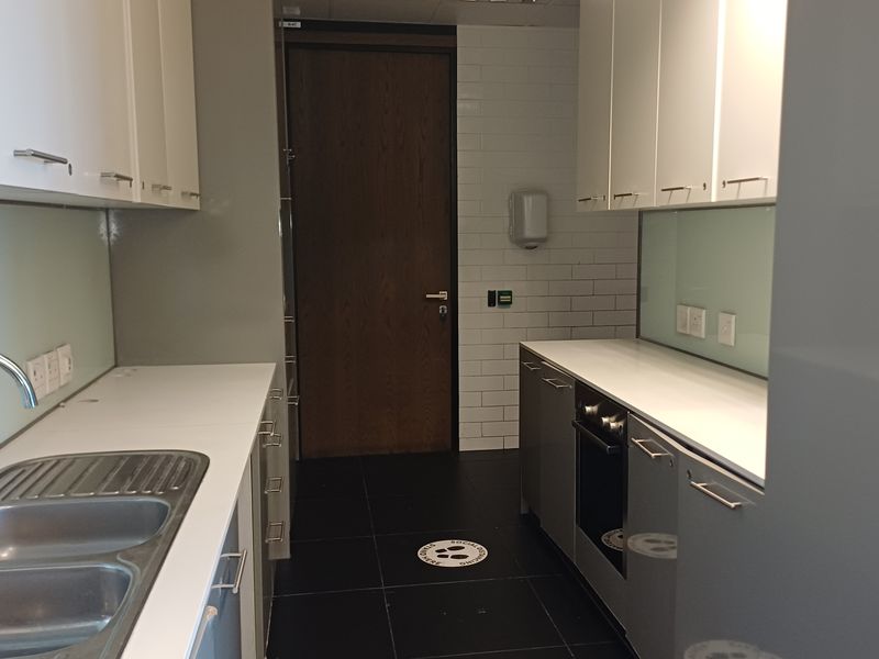A clean, modern office kitchenette featuring built-in cabinetry, a stainless steel sink, a built-in oven, and a glass splashback with tiled wall accents.