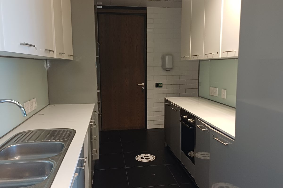 A clean, modern office kitchenette featuring built-in cabinetry, a stainless steel sink, a built-in oven, and a glass splashback with tiled wall accents.