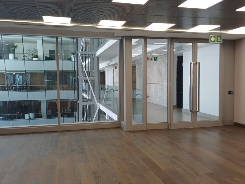 A modern, empty office space featuring high-quality timber flooring, recessed ceiling lights, and large glass partitions overlooking an internal atrium.