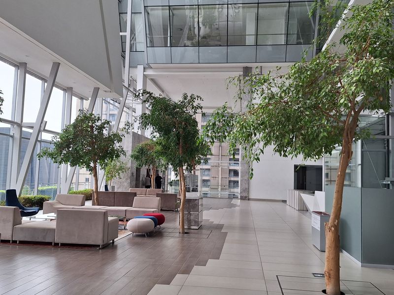 Spacious modern office atrium featuring high ceilings, floor-to-ceiling glass windows, integrated indoor landscaping, and polished tile flooring in a professional corporate environment.