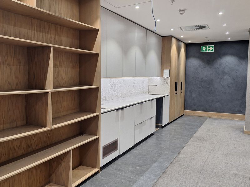 Modern office kitchenette area featuring built-in wooden shelving, sleek cabinetry with stone countertops, and integrated sink, finished with professional recessed lighting.
