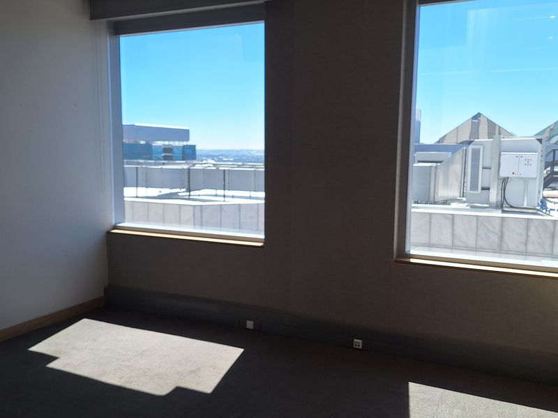 A bright, empty office space featuring large windows with city views, recessed ceiling lighting, and neutral carpeted flooring.