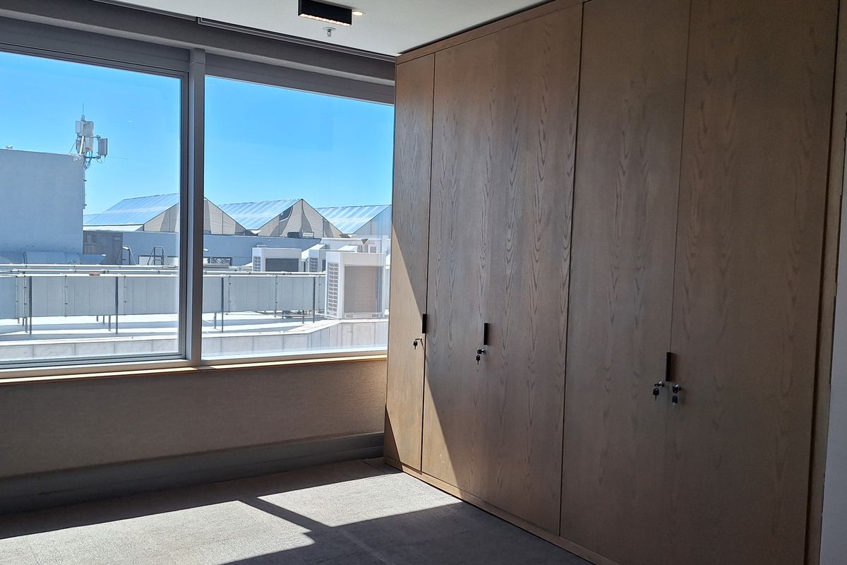 A bright, empty office space featuring extensive built-in wooden storage cabinetry, large windows with natural light, and modern recessed ceiling lighting.
